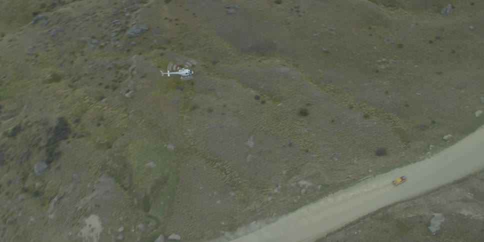 A helicopter flies over and follows a truck speeding on a desert dirt road kicking up dust