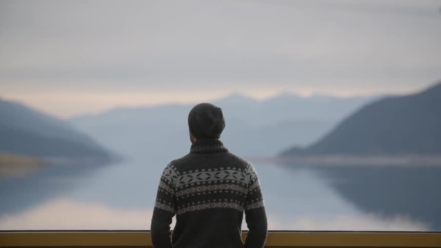 A man is standing by the shore overlooking a mountain range past the water