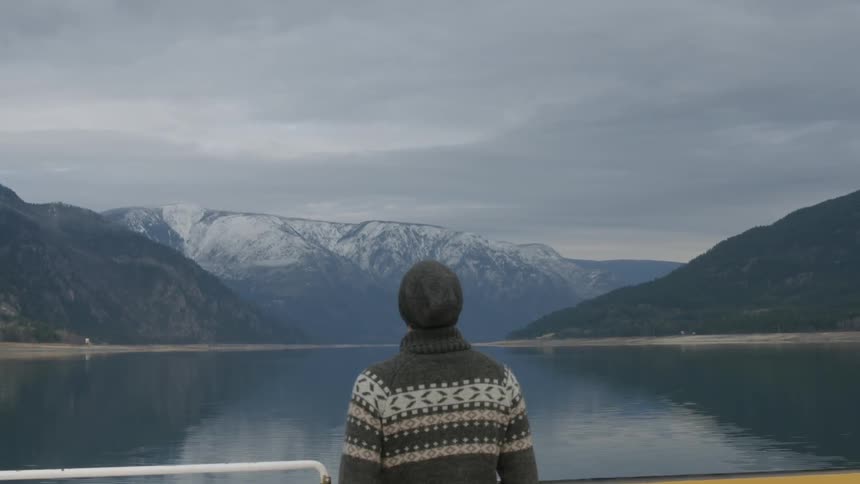 A man looking out over a lake surrounded by snow-capped mountains on a cloudy day