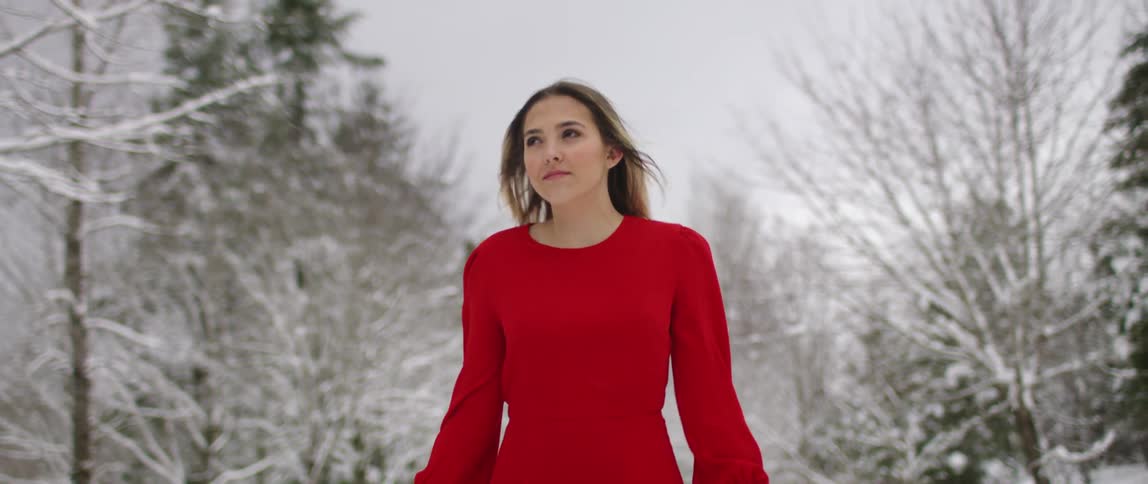Woman in a red dress walks through snow covered forest in a windy day