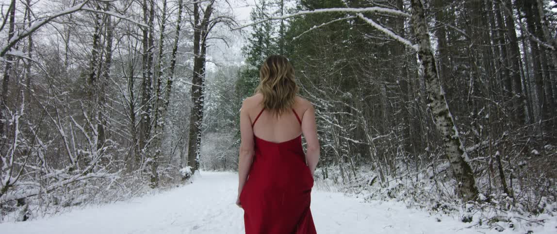 A woman in a red dress walking in the forest as it is snowing