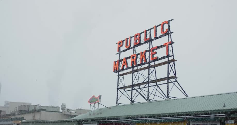 Snow falls on a rooftop with a neon public market sign glowing on top
