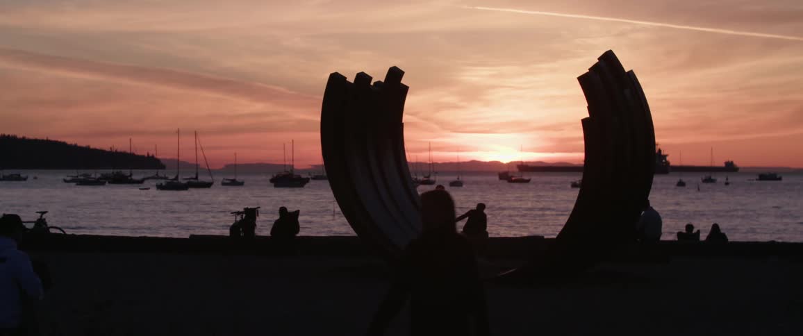 People walking and sitting near the shore with boats and the sunset in the background