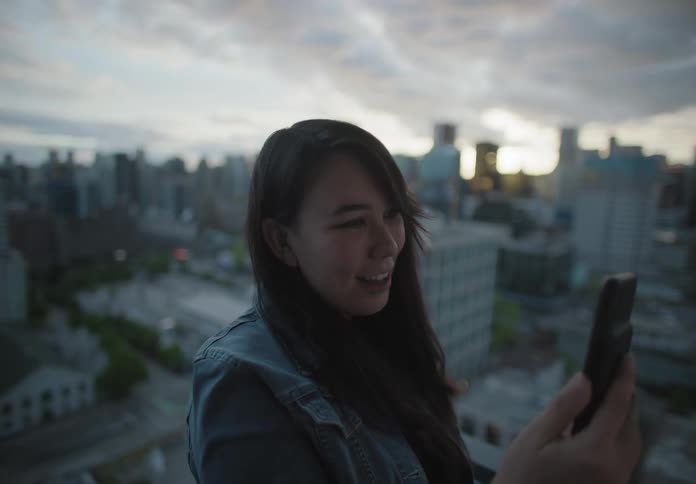 Woman standing on balcony answers video call on smartphone at sunset