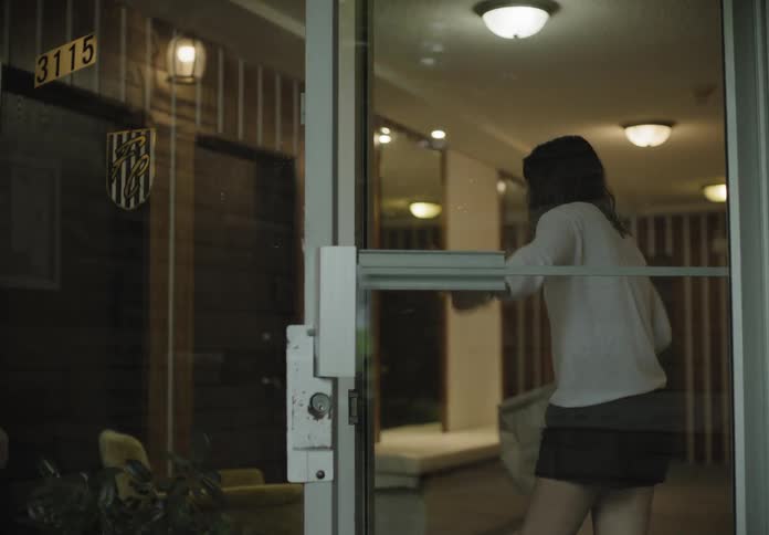 Female casually walks to the glass entry door to the apartment building as she waves to the delivery man then opens the door to accept the brown paper bag and turns and walks back through the building