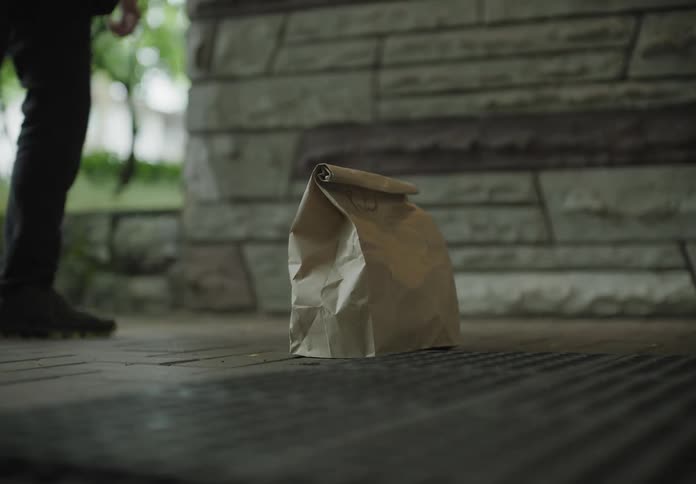 A man delivers a paper bag to a home leaving it at the front door and stepping away as a woman opens the door and picks it up