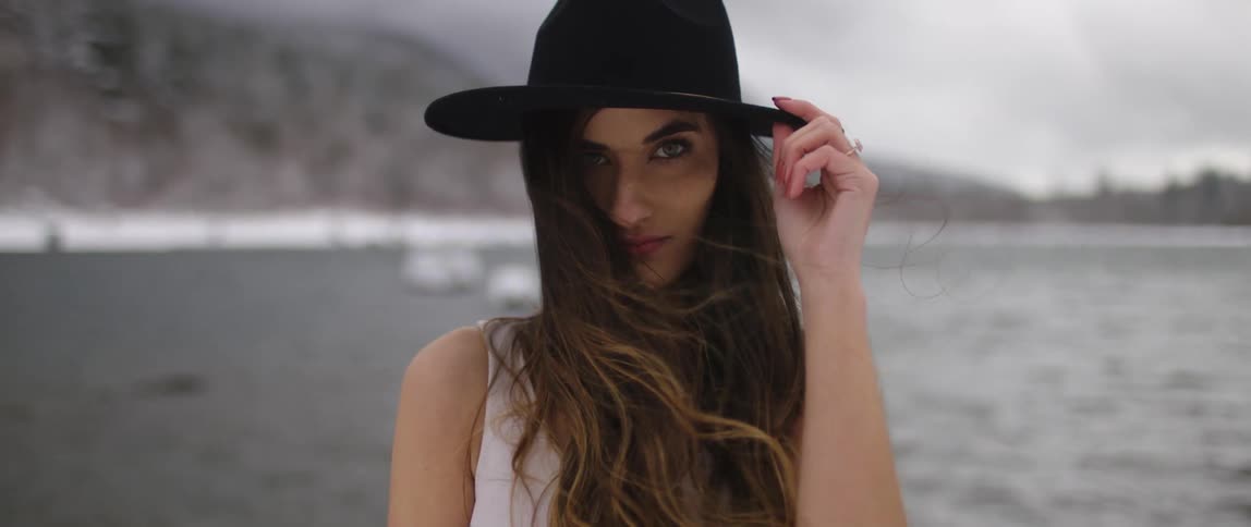 A white woman with a hat stands by a body of water smiling
