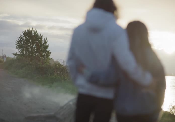 A couple stands near the water with their arms around each other at sunset