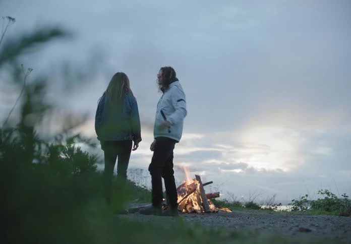 A man lighting a bonfire before walking over to a woman and wrapping his arm around her shoulders