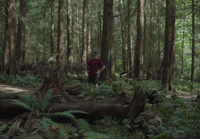 A man jogging through the woods stops to check his cell phone and another man jogs past him