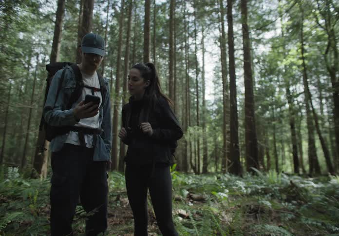 A man and woman hike through a forest and then stop and look at his cell phone and turn around