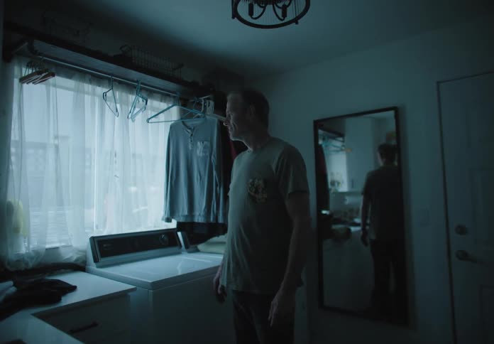A man looking out the window while standing in a dim room before turning to look into the camera