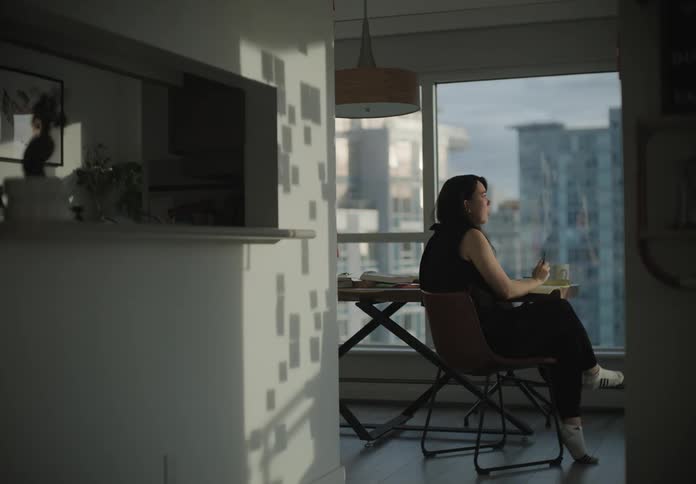 A woman sits at her table in front of large windows overlooking the city drinking coffee and writing