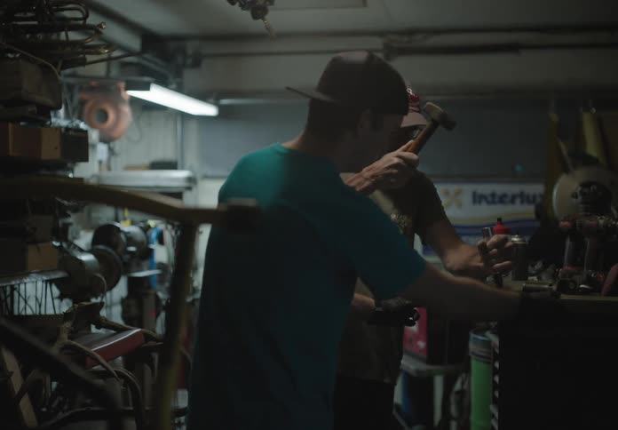 Two men in a workshop one holds a piece of wood while the other uses tools on it
