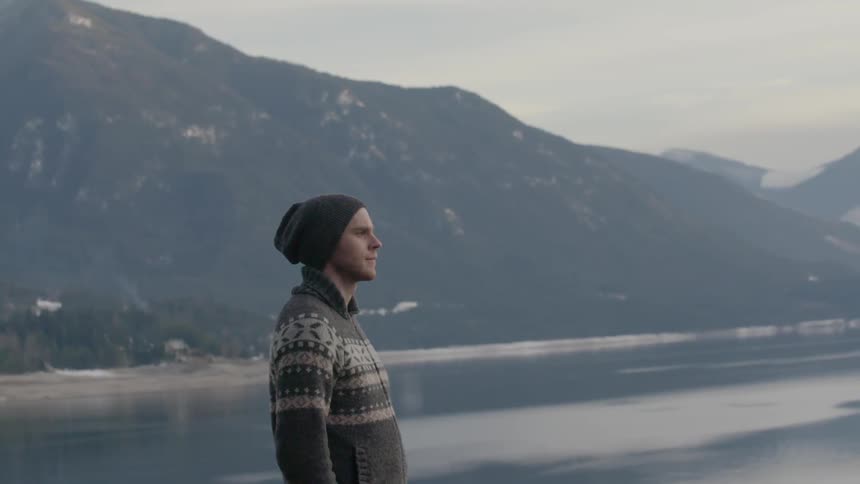 A man stands staring from an elevated position near a lake and mountains