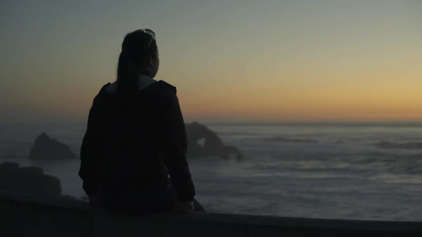 Woman sitting on wall watching golden sunset with peaceful reflective mood.