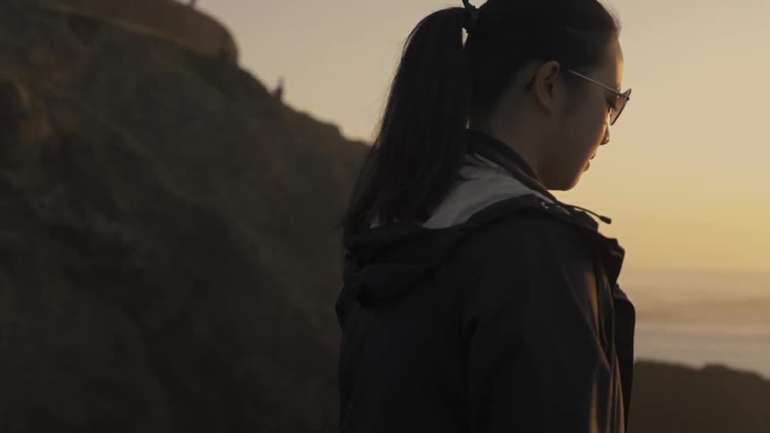 A woman walks a rocky ledge overlooking the ocean at sunset