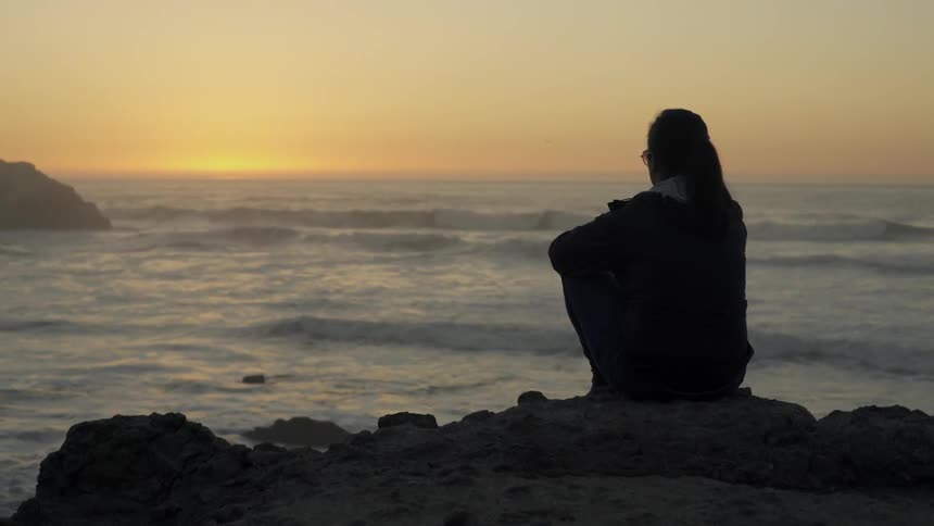 A woman sitting on an ocean-side cliff at sunset