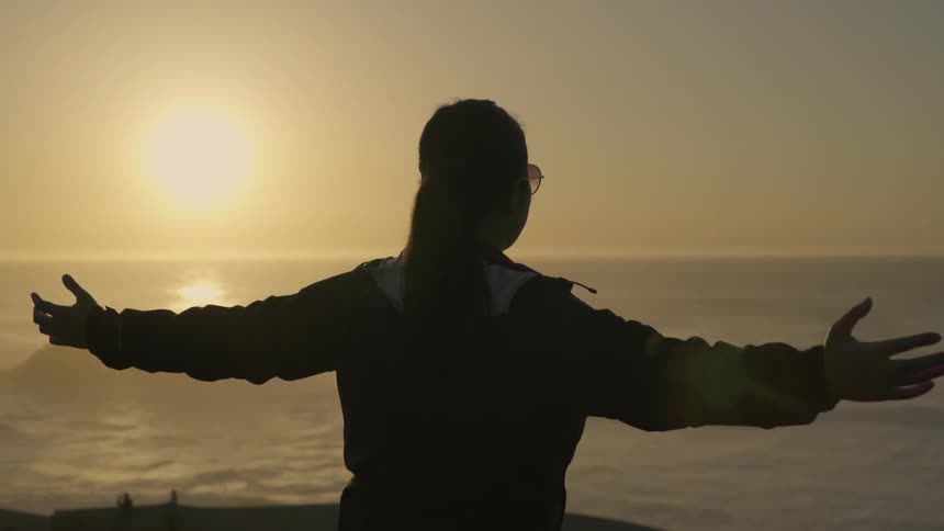 A woman standing with arms outstretched above a shore as the sun sets