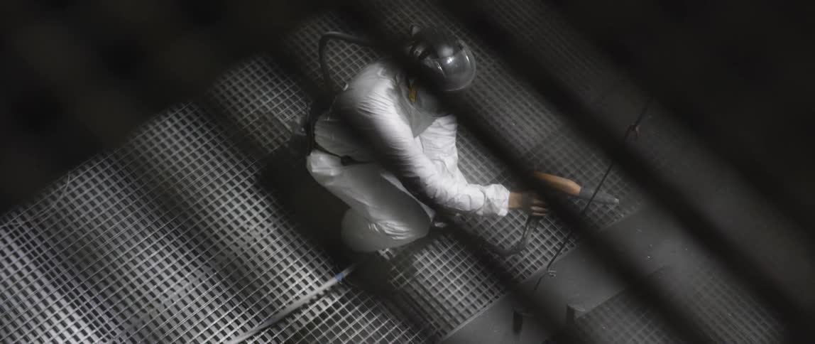 A man wearing coveralls and a face shield uses a power sprayer to paint and coat a suspended piece of metal