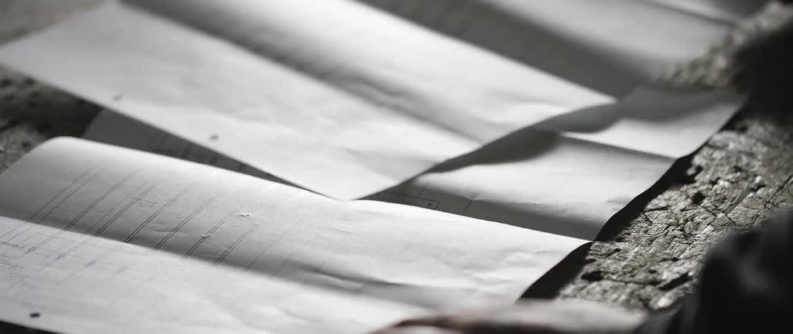 A man places paper plans down on a wooden worktop that is scratched