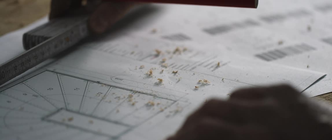 A craftsman moves the ruler to the edge of a plan on paper which is dotted with sawdust