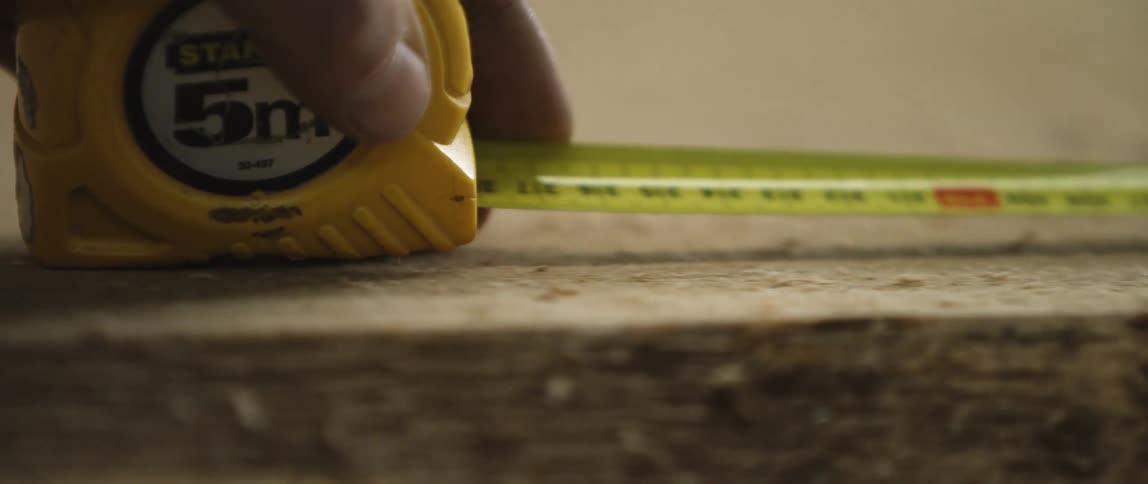 A white man measures a length of wood with a tape measure