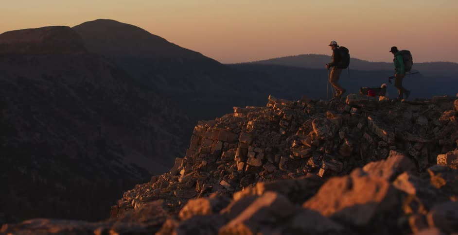 A pair of hikers and a dog walking towards a rocky cliff at sunset