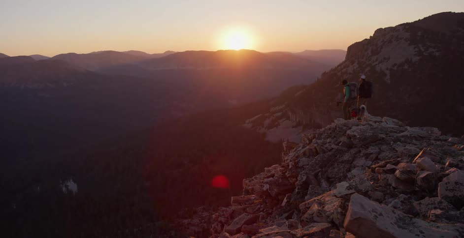 Two people and a dog hiking on a cliff with mountains and the sky in the background