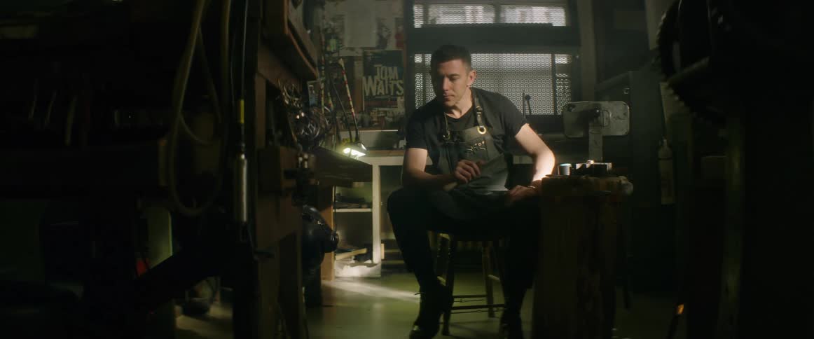 A man in a safety apron sitting on a stool in a workshop while lost in thought