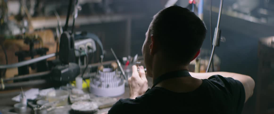 A man sits at a table in a workshop using tools and working on something