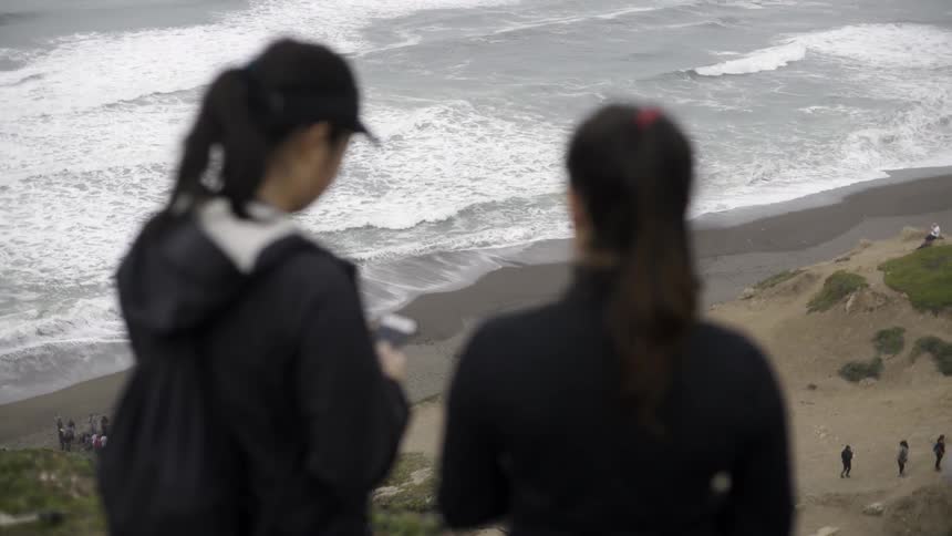 Two women are standing on the top of a hill photographing the beach below