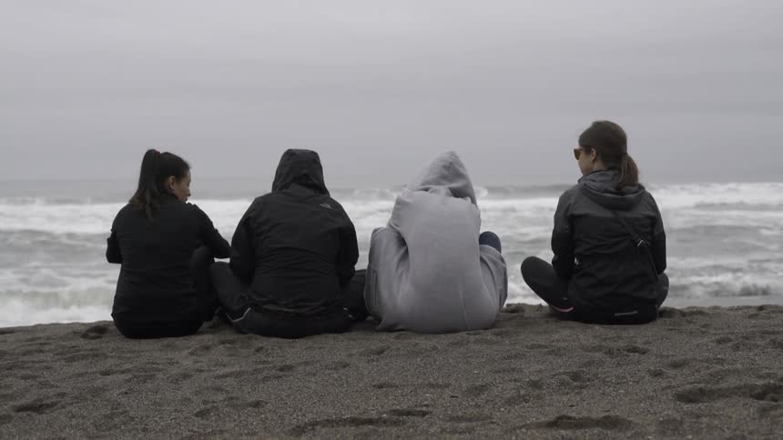 Four women sit on a beach facing the ocean on a cloudy day