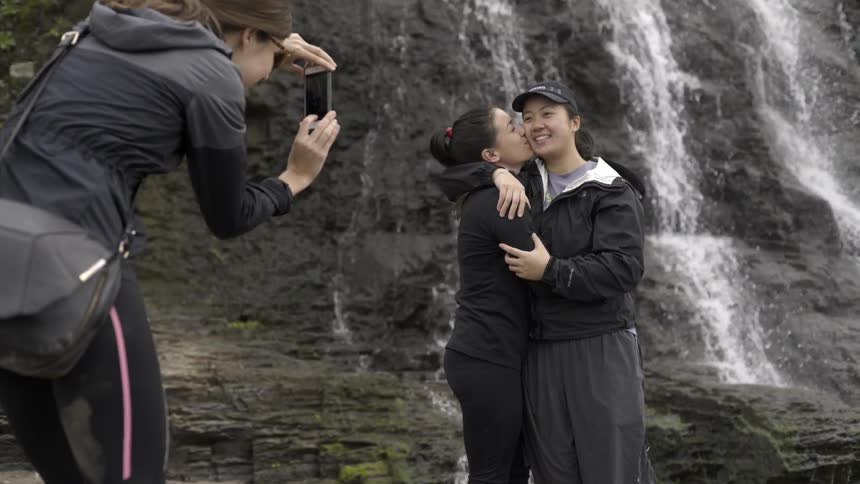 A young woman kissing another woman's cheek as the are photographed in front of a waterfall