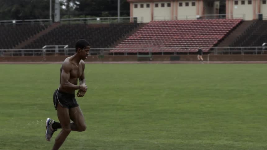 Shirtless man jogging across a sports field in a sprinkling rain
