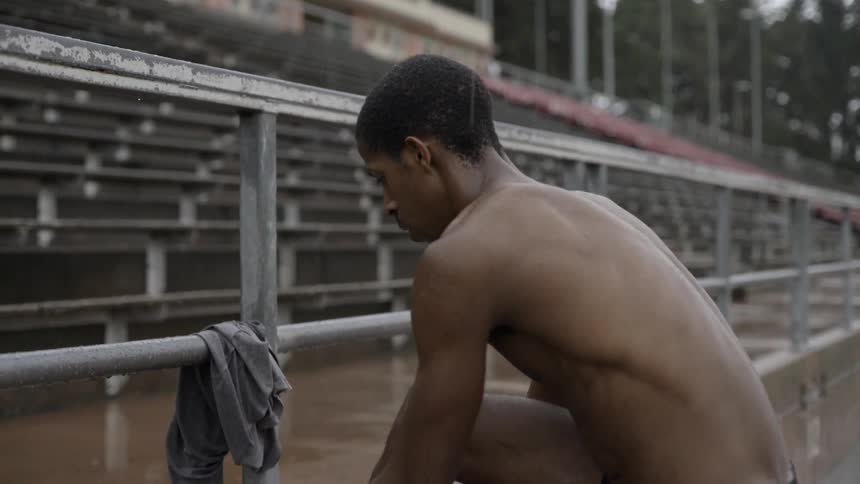 A shirtless man ties his shoelaces at an outdoor racing track on a rainy day