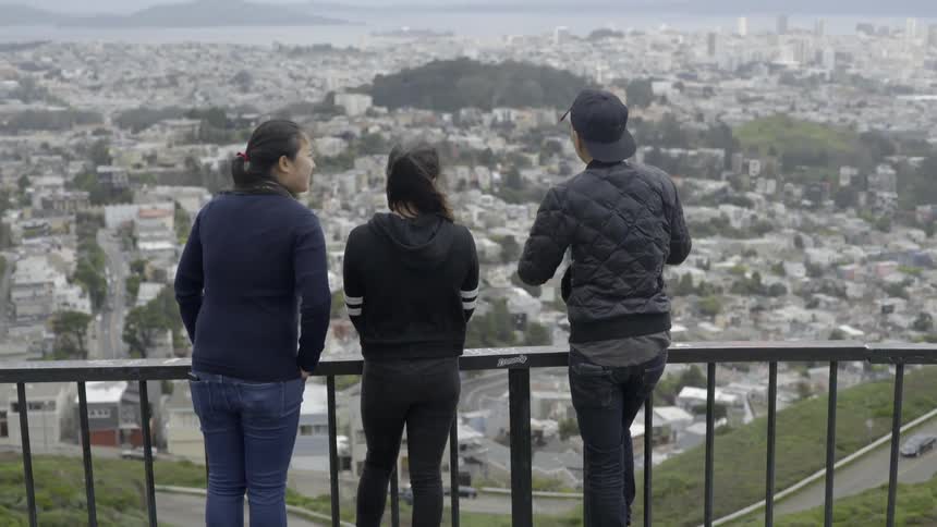 Three people are standing near a railing above a town on a cloudy day