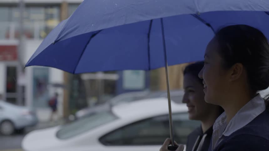 Two women walk down rainy street under umbrella with joyful friendship energy.