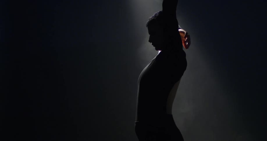 Dancer poses and turns under dim spotlight in dark shadowed room