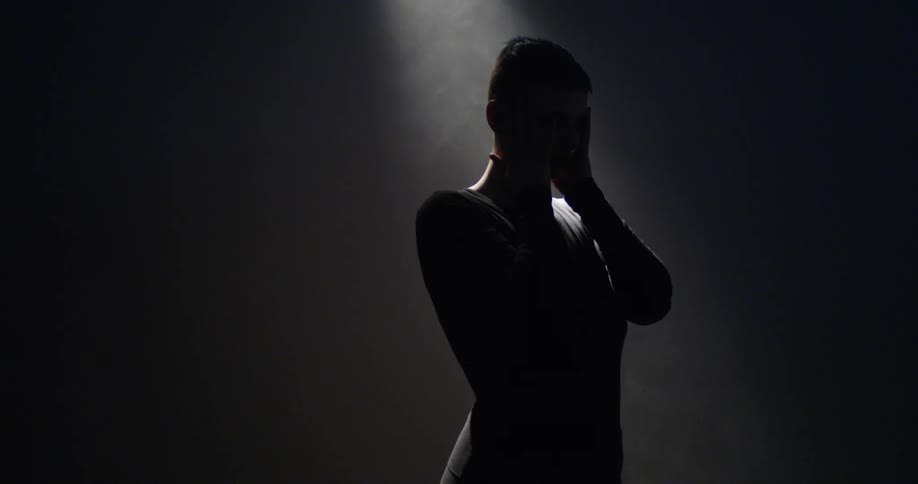 A woman stands with a light shining down on her touching her head and face with both hands