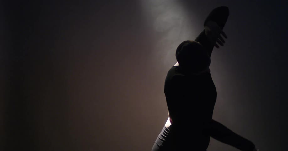 A white male dancer moves slowly in light and shadow