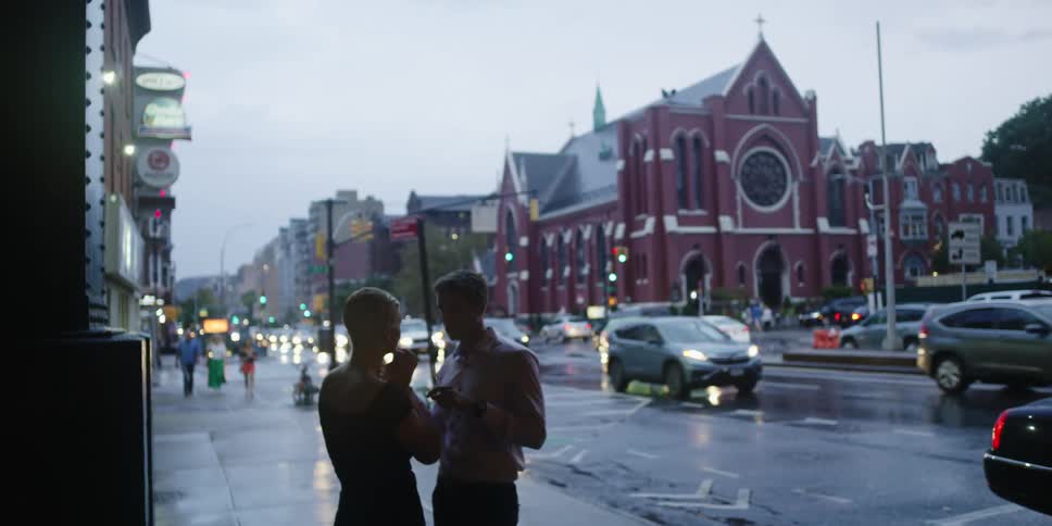 A couple stand on the sidewalk talking as traffic goes by on the street