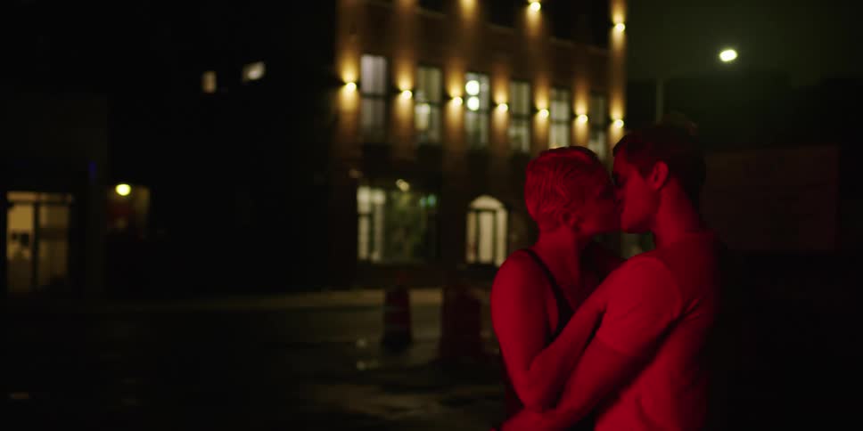 A man and woman make out on a city sidewalk at night with a reddish light glow
