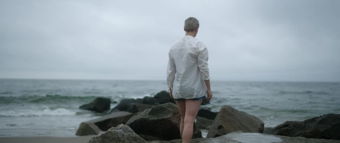 A woman wearing shorts and a white shirt walks on the rocks as the waves break and come to shore