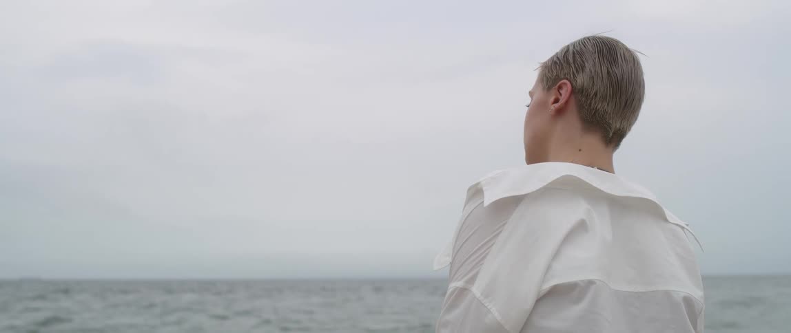 A woman stands in front of the water and looks out