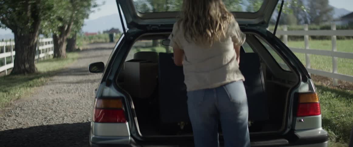 Woman loads luggage in car on gravel road between white fenced pastures