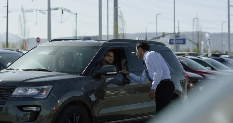 A car salesman approaches a couple in a vehicle on the car lot and interacts with them