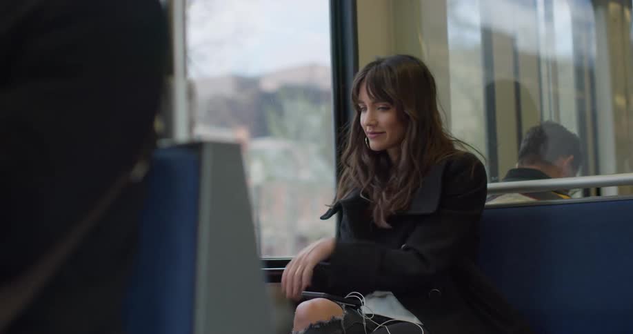 A woman looks out the window of a bus and shakes a pair of earbuds in her hands