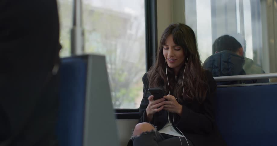 A woman sits on a city bus listening to ear buds connected to her smart phone