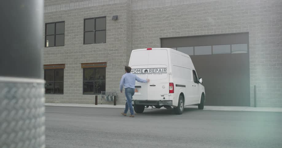 A man exits the back of his work van into a parking lot outside of a large building